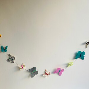 K&ouml;nnte beinhalten: Eine Girlande aus bunten Papier-Schmetterlingen, mit verschiedenen Mustern und Farben, darunter Gelb, Blau, Gr&uuml;n, Rosa und Wei&szlig;, ist an einer wei&szlig;en Wand befestigt.