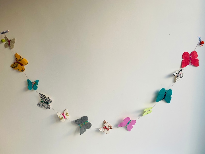 K&ouml;nnte beinhalten: Eine Girlande aus bunten Papier-Schmetterlingen, mit verschiedenen Mustern und Farben, darunter Gelb, Blau, Gr&uuml;n, Rosa und Wei&szlig;, ist an einer wei&szlig;en Wand befestigt.