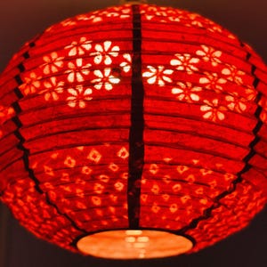 May include: A spherical red paper lantern with a floral and geometric pattern. The lantern is lit from within, producing a warm glow. The design includes white flower cutouts and diamond shapes.