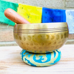 May include: A gold singing bowl with a wooden striker, resting on a teal cushion with gold embroidery. The bowl has a hammered texture and two bands around the rim.