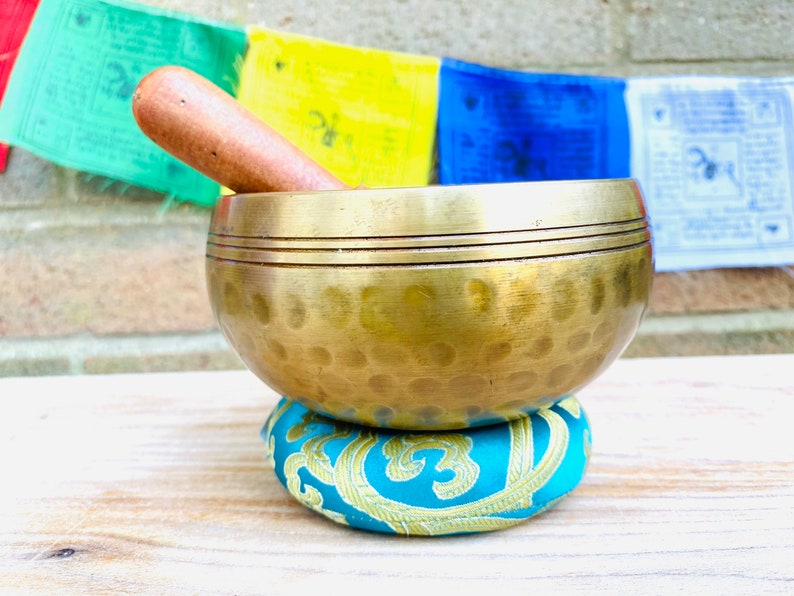 May include: A gold singing bowl with a wooden striker, resting on a teal cushion with gold embroidery. The bowl has a hammered texture and two bands around the rim.