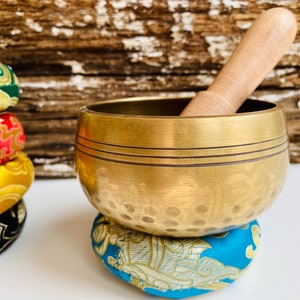 May include: A gold singing bowl with a wooden striker, resting on a blue and gold patterned cushion. The bowl has a hammered texture and a decorative design.