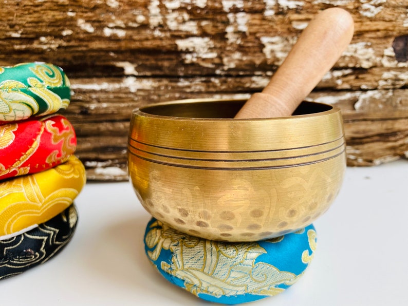 May include: A gold singing bowl with a wooden striker, resting on a blue and gold patterned cushion. The bowl has a hammered texture and a decorative design.