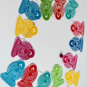 K&ouml;nnte beinhalten: Eine bunte Girlande aus Papierschnitten, die Buddha-Figuren in verschiedenen Posen darstellen. Jede Ausschneidung hat eine andere Farbe, darunter Rot, Gelb, Gr&uuml;n, Blau und Lila. Die Schnur ist mit kleinen Perlen besetzt.