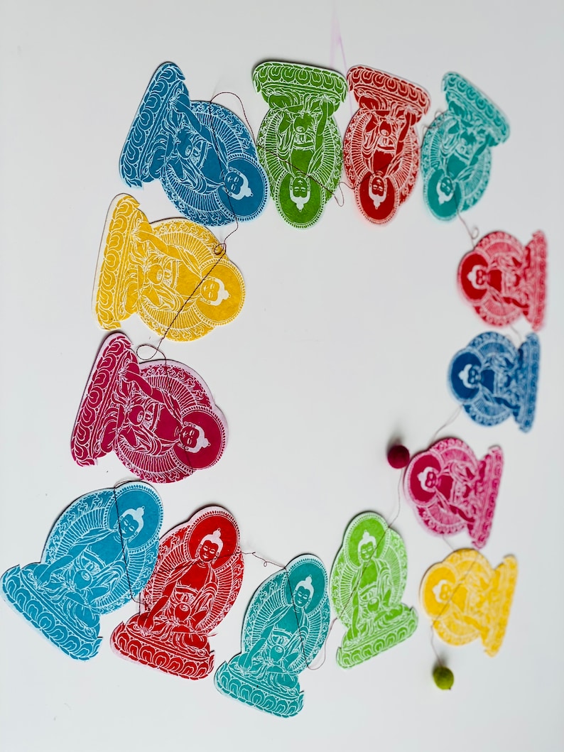 K&ouml;nnte beinhalten: Eine bunte Girlande aus Papierschnitten, die Buddha-Figuren in verschiedenen Posen darstellen. Jede Ausschneidung hat eine andere Farbe, darunter Rot, Gelb, Gr&uuml;n, Blau und Lila. Die Schnur ist mit kleinen Perlen besetzt.