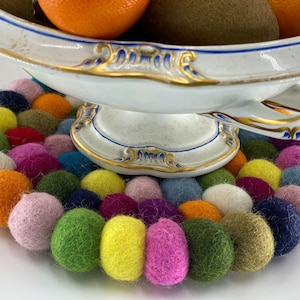 May include: A white porcelain bowl with gold trim sits on a colorful felt coaster. The coaster is made of many small, round felt balls in various colors, including pink, green, yellow, blue, and orange.
