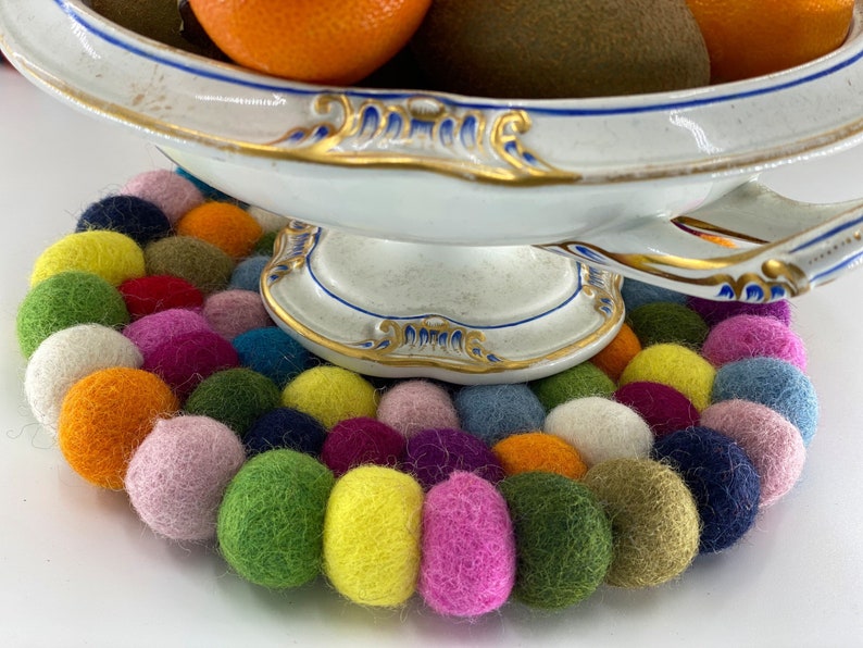 May include: A white porcelain bowl with gold trim sits on a colorful felt coaster. The coaster is made of many small, round felt balls in various colors, including pink, green, yellow, blue, and orange.
