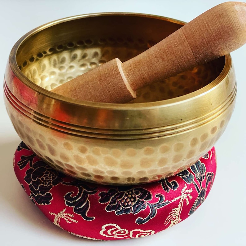 May include: A gold singing bowl with a wooden striker, resting on a red and black floral cushion.