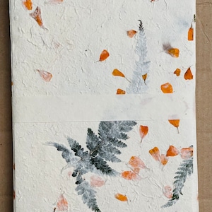 May include: A white handmade paper journal with pressed orange flower petals and green ferns. The journal has a textured surface and a white ribbon closure.