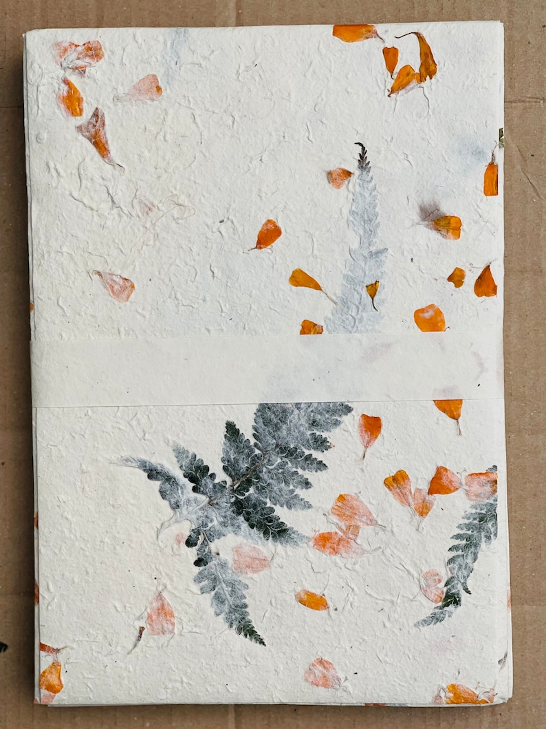 May include: A white handmade paper journal with pressed orange flower petals and green ferns. The journal has a textured surface and a white ribbon closure.
