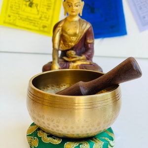 May include: A brass singing bowl with a wooden striker, resting on a green and gold cushion. The bowl has a hammered texture and a double rim. A golden Buddha statue sits in the background.