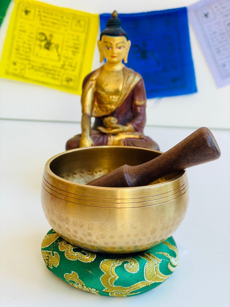 May include: A brass singing bowl with a wooden striker, resting on a green and gold cushion. The bowl has a hammered texture and a double rim. A golden Buddha statue sits in the background.
