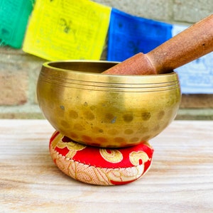 May include: A brass singing bowl with a red and gold patterned cushion. The bowl has a hammered finish and is resting on a wooden surface. A wooden striker is resting on top of the bowl.