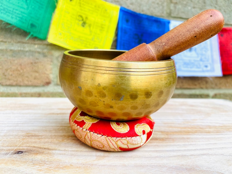 May include: A brass singing bowl with a red and gold patterned cushion. The bowl has a hammered finish and is resting on a wooden surface. A wooden striker is resting on top of the bowl.