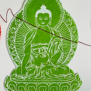 K&ouml;nnte beinhalten: Gr&uuml;ner Papierschnitt eines Buddha, der mit einer Schale auf dem Scho&szlig; in Meditation sitzt. Der Buddha ist von einem Heiligenschein und einem Lotusblumenmuster umgeben.