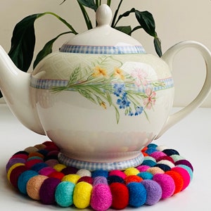 May include: A white teapot with a floral design sits on a colorful felt coaster. The teapot has a blue and white checkered trim and a floral pattern with pink, blue, and yellow flowers.