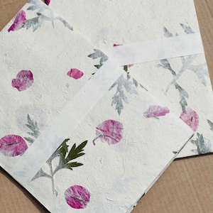 May include: Two white paper cards with pressed flowers and leaves. The cards are blank and have a textured surface. The flowers are pink and the leaves are green.