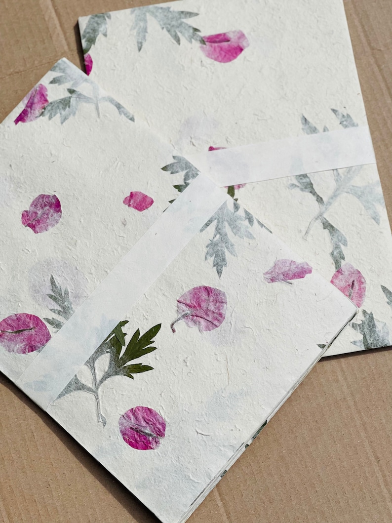 May include: Two white paper cards with pressed flowers and leaves. The cards are blank and have a textured surface. The flowers are pink and the leaves are green.
