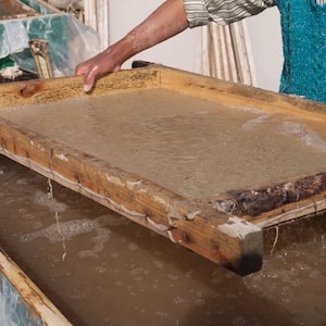 May include: A person lifts a wooden frame out of a basin of water. The frame is filled with a thin layer of pulp, which is used to make paper.