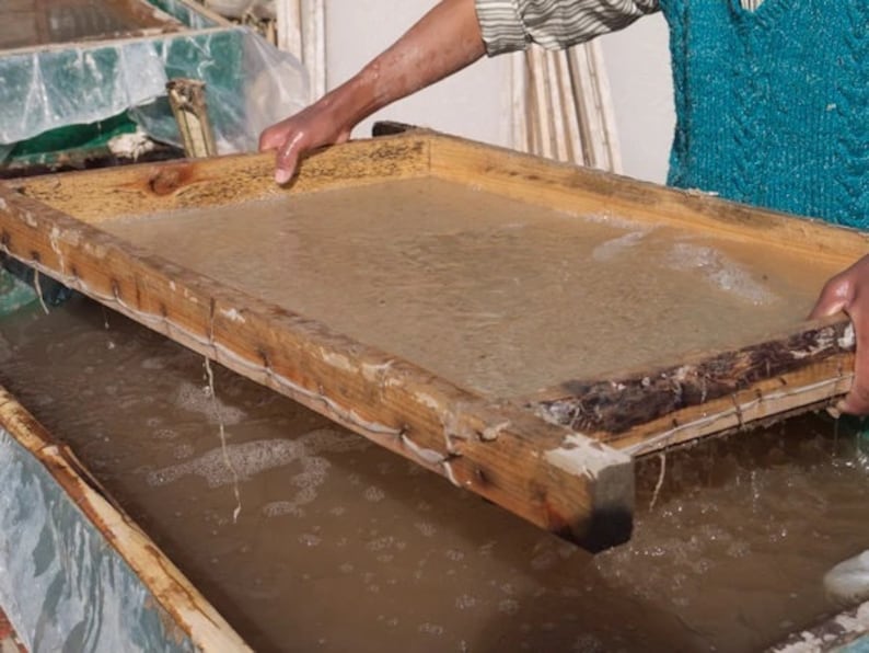 May include: A person lifts a wooden frame out of a basin of water. The frame is filled with a thin layer of pulp, which is used to make paper.