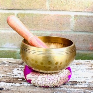 May include: A gold singing bowl with a wooden striker, resting on a purple and gold cushion. The bowl has a hammered texture and a wide rim.