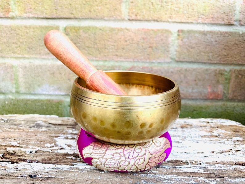 May include: A gold singing bowl with a wooden striker, resting on a purple and gold cushion. The bowl has a hammered texture and a wide rim.