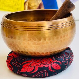 May include: A golden singing bowl with a wooden striker, resting on a red and black patterned cushion.