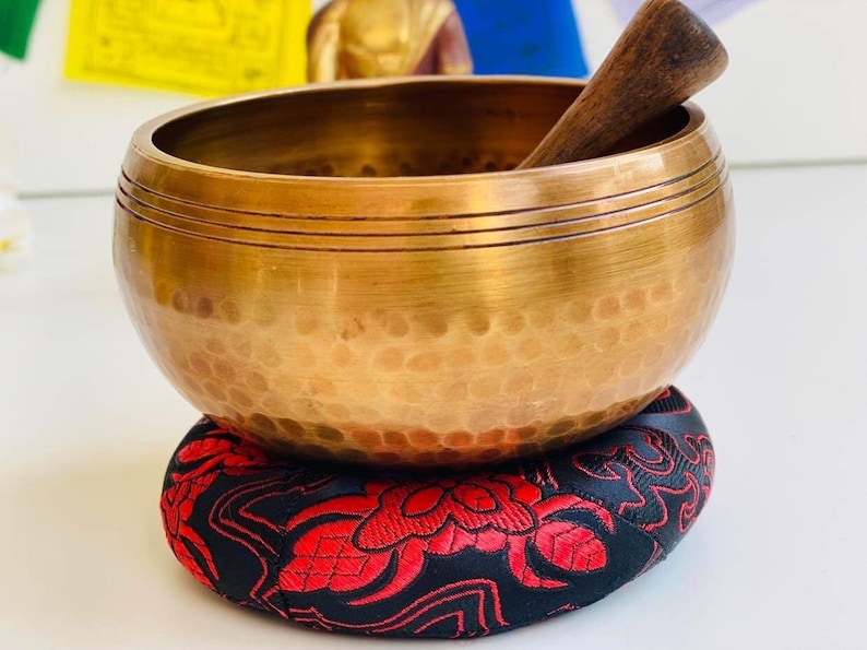 May include: A golden singing bowl with a wooden striker, resting on a red and black patterned cushion.