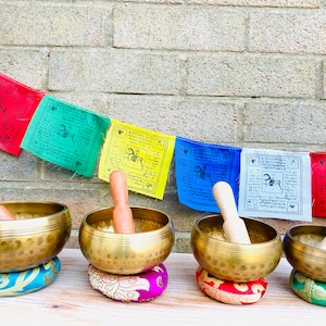 May include: Four antique brass singing bowls with wooden strikers, each resting on a colorful embroidered cushion. The bowls are arranged in a row on a wooden surface, with prayer flags hanging in the background.