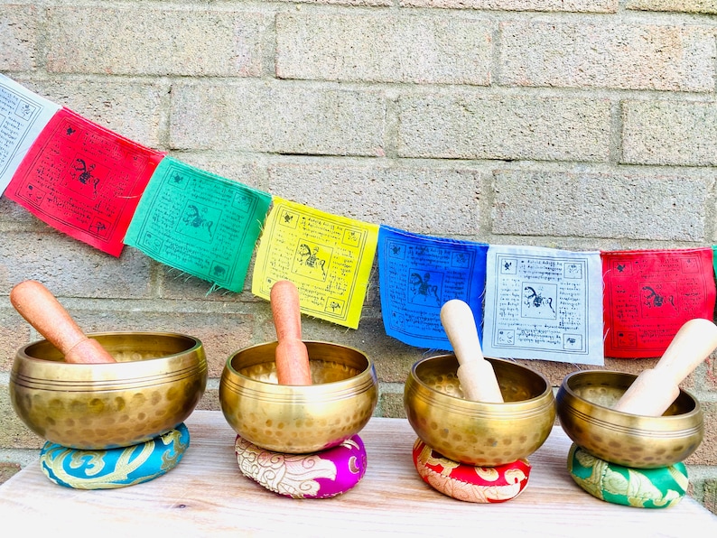 May include: Four antique brass singing bowls with wooden strikers, each resting on a colorful embroidered cushion. The bowls are arranged in a row on a wooden surface, with prayer flags hanging in the background.
