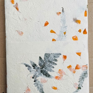 May include: A white handmade paper journal with pressed orange flower petals and green ferns. The journal is bound with a white ribbon.