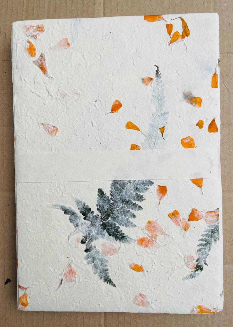 May include: A white handmade paper journal with pressed orange flower petals and green ferns. The journal is bound with a white ribbon.