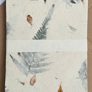 May include: A white handmade paper journal with pressed leaves and flowers. The paper has a textured, organic look. The journal has a white ribbon bookmark.