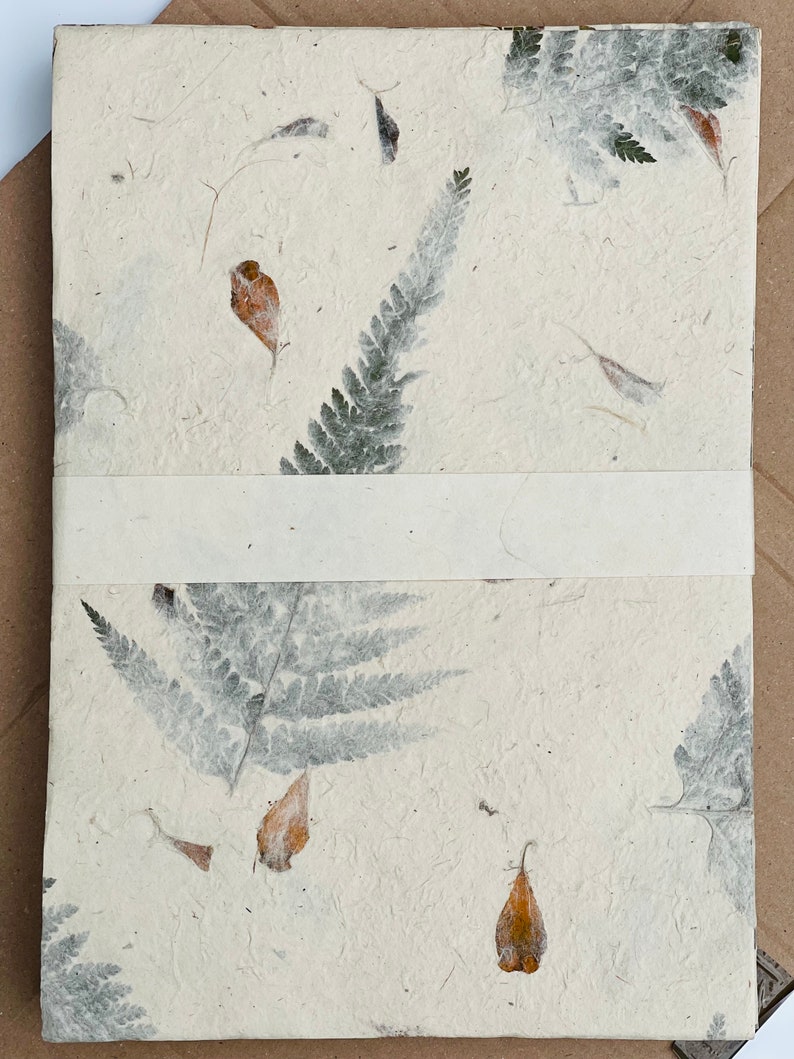 May include: A white handmade paper journal with pressed leaves and flowers. The paper has a textured, organic look. The journal has a white ribbon bookmark.