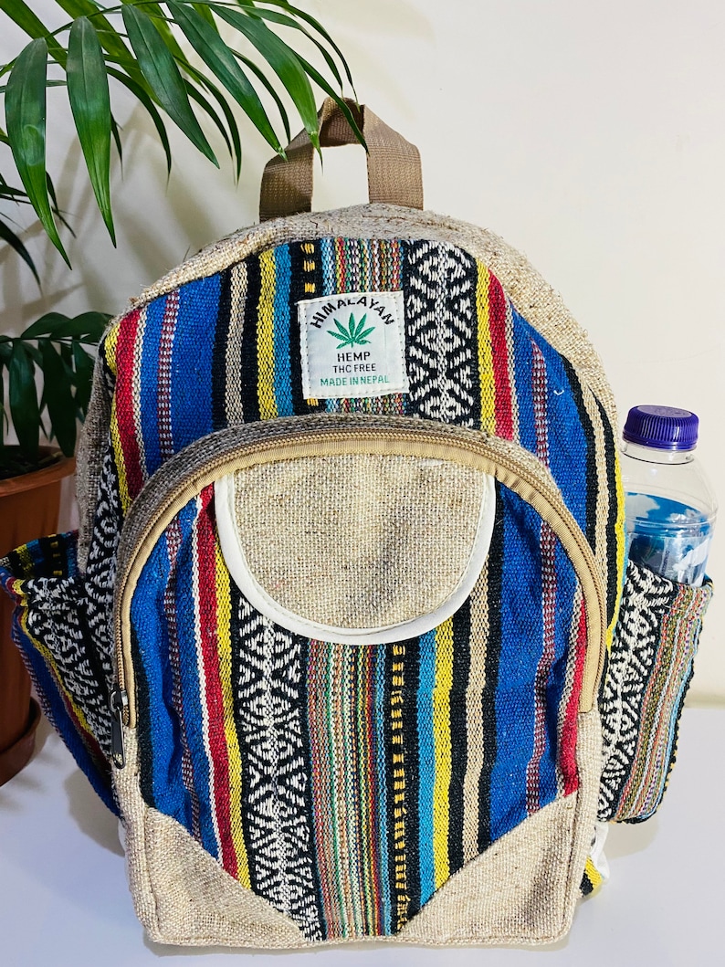 May include: A multi-colored striped hemp backpack with a brown leather strap and a small pocket on the side. The backpack has a label that reads "Himalayan Hemp THC Free Made in Nepal".