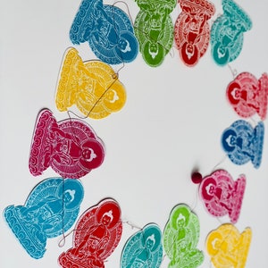 K&ouml;nnte beinhalten: Eine Girlande aus bunten Papierschnitten, die Buddha-Figuren in sitzender Meditationshaltung darstellen. Jede Ausschneidung hat eine andere Farbe, darunter Gelb, Gr&uuml;n, Rot, Blau und Orange. Die Ausschnitte sind mit einem d&uuml;nnen Faden aneinandergereiht.