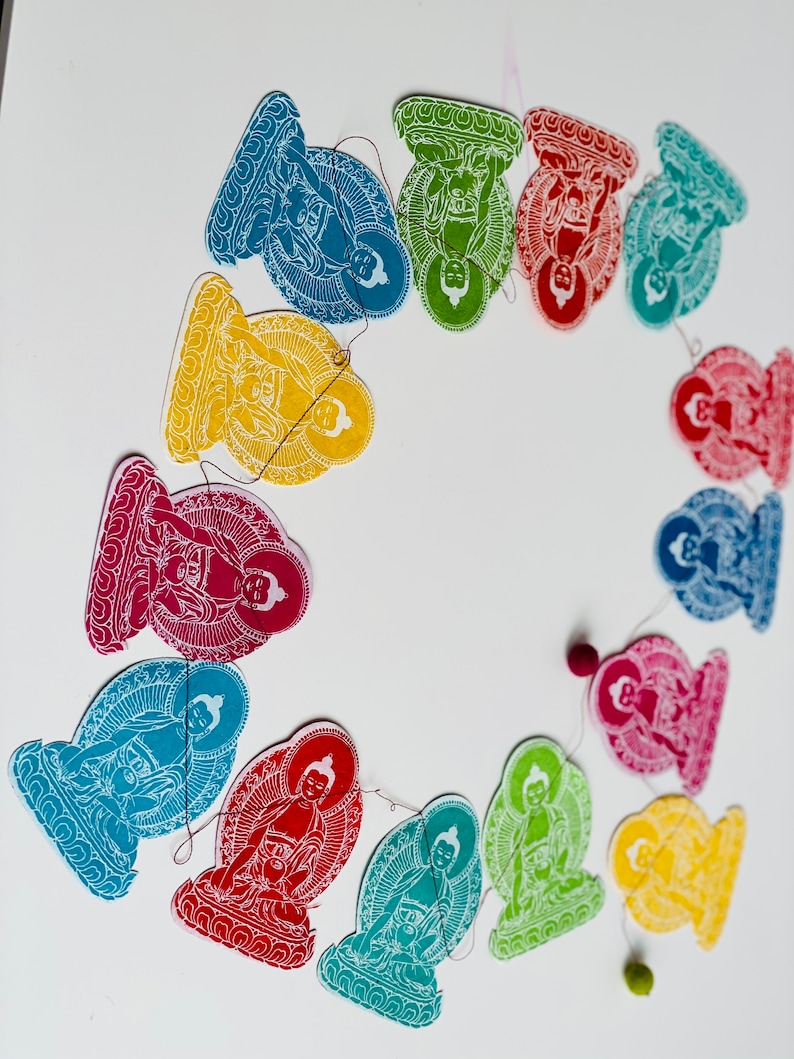 K&ouml;nnte beinhalten: Eine Girlande aus bunten Papierschnitten, die Buddha-Figuren in sitzender Meditationshaltung darstellen. Jede Ausschneidung hat eine andere Farbe, darunter Gelb, Gr&uuml;n, Rot, Blau und Orange. Die Ausschnitte sind mit einem d&uuml;nnen Faden aneinandergereiht.