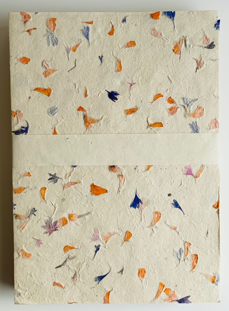 May include: A handmade notebook with a cream-colored cover featuring a unique design of pressed orange, blue, and pink flower petals.