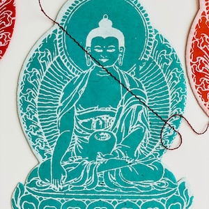 K&ouml;nnte beinhalten: Ein blaugr&uuml;ner Papierschnitt eines sitzenden Buddha mit einem wei&szlig;en Heiligenschein und wei&szlig;en Details. Der Buddha sitzt in meditativer Haltung mit den H&auml;nden im Scho&szlig;.