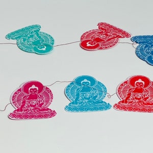 K&ouml;nnte beinhalten: Eine Girlande aus bunten Papierschnitten von Buddha-Figuren, jede mit einer anderen Farbe und einem anderen Design. Die Figuren sind durch eine d&uuml;nne Schnur verbunden und h&auml;ngen an einem wei&szlig;en Hintergrund.