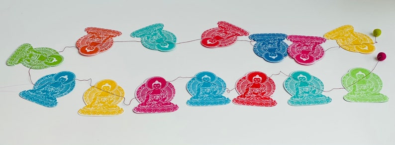 K&ouml;nnte beinhalten: Eine Girlande aus bunten Papierschnitten von Buddha-Figuren, jede mit einer anderen Farbe und einem anderen Design. Die Figuren sind durch eine d&uuml;nne Schnur verbunden und h&auml;ngen an einem wei&szlig;en Hintergrund.