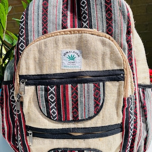 May include: A beige hemp backpack with black and red striped fabric accents. The backpack has two large zipped pockets and a smaller pocket on the front. The front pocket has a label that reads "Himalayan Hemp THC Free Nepalese Hemp".