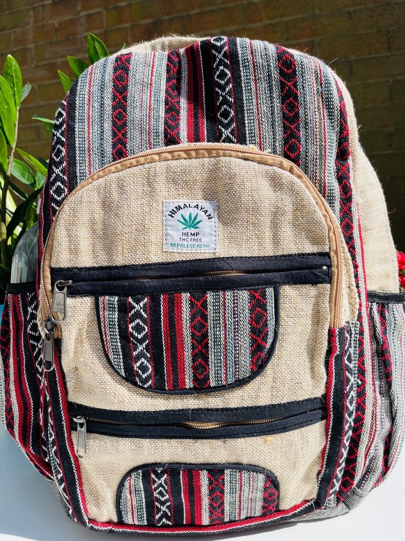 May include: A beige hemp backpack with black and red striped fabric accents. The backpack has two large zipped pockets and a smaller pocket on the front. The front pocket has a label that reads "Himalayan Hemp THC Free Nepalese Hemp".