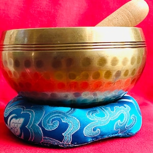 May include: A gold singing bowl with a wooden striker, resting on a blue patterned cushion. The bowl has a hammered surface and a slightly flared lip.