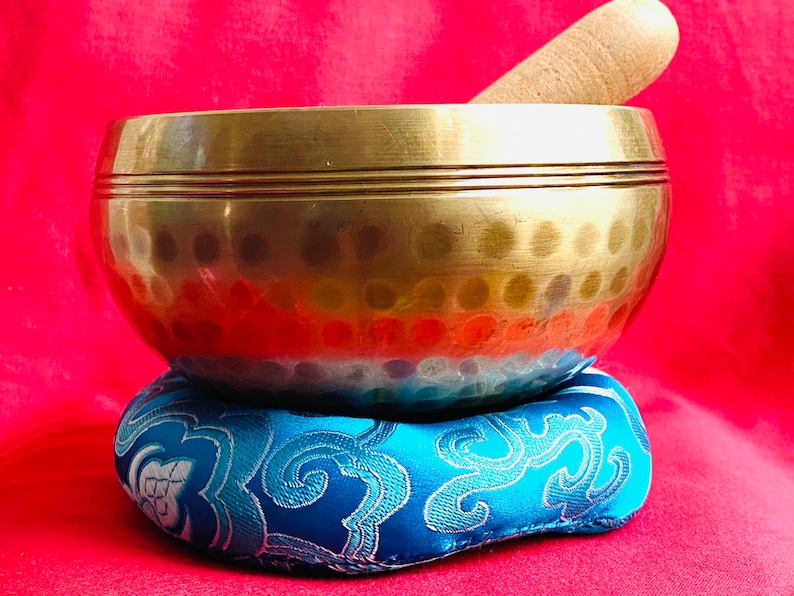 May include: A gold singing bowl with a wooden striker, resting on a blue patterned cushion. The bowl has a hammered surface and a slightly flared lip.