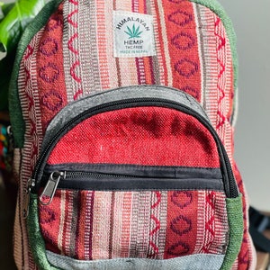 May include: A red, green, and white striped hemp backpack with a black zipper and a small pocket on the front. The backpack has a label that says "Himalayan Hemp THC Free Made in Nepal".