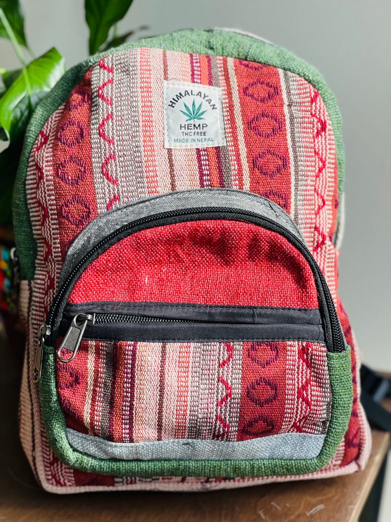 May include: A red, green, and white striped hemp backpack with a black zipper and a small pocket on the front. The backpack has a label that says "Himalayan Hemp THC Free Made in Nepal".