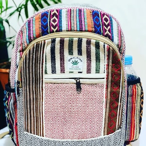Op de afbeelding: Multicolor gestreepte hennep rugzak met een groot voorvak en een kleiner vak met rits. De rugzak heeft een bruine leren rits trek en een label met de tekst "Himalayan THC Free Made in Nepal".