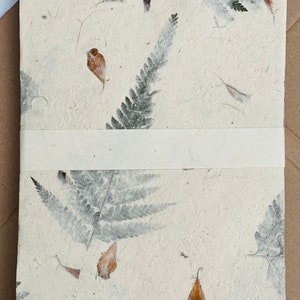 May include: A white handmade paper journal with pressed leaves and flowers. The paper has a textured, organic look. The journal has a white ribbon bookmark.