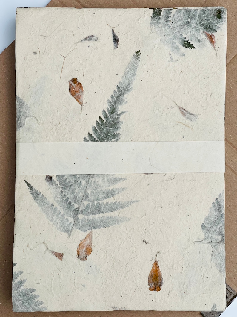 May include: A white handmade paper journal with pressed leaves and flowers. The paper has a textured, organic look. The journal has a white ribbon bookmark.